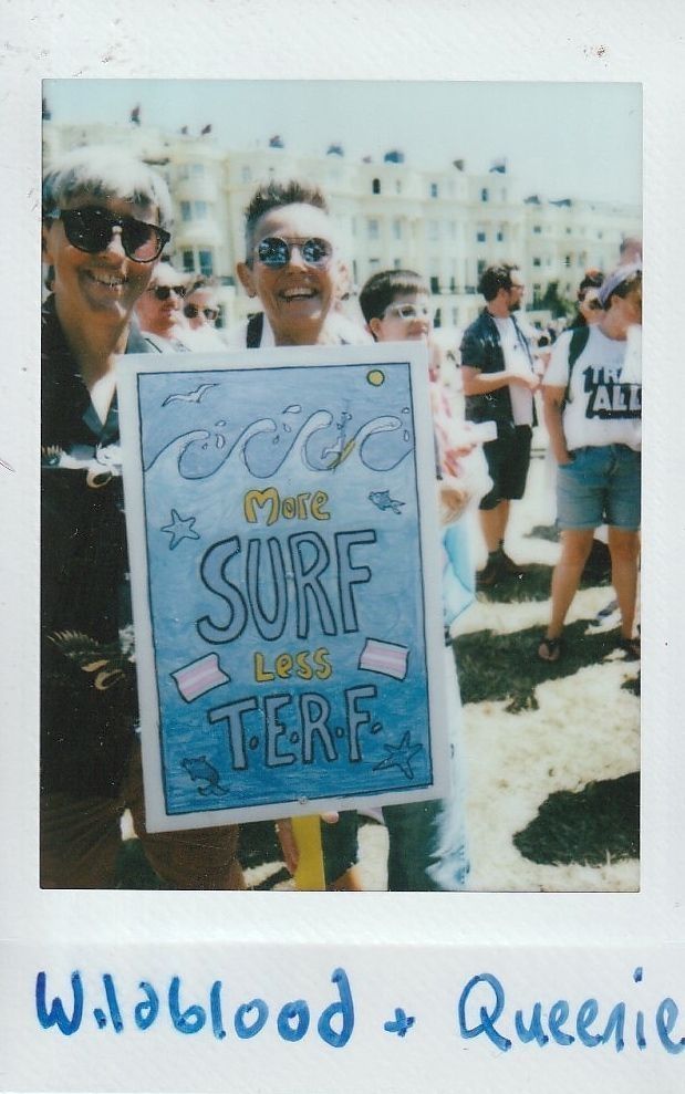 People are smiling outdoors, holding a sign reading "More Surf Less TERF" during a sunny day.