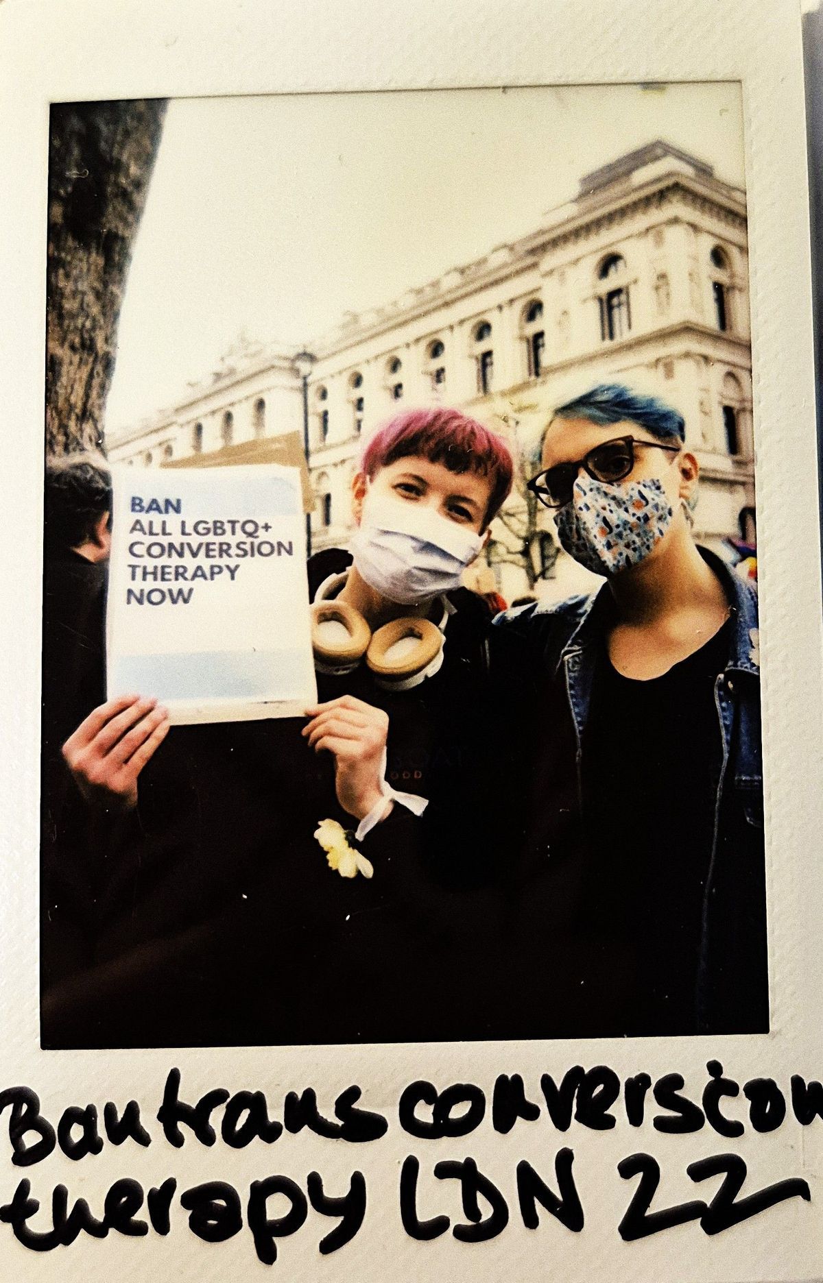 Two people wearing masks hold a sign that reads, "Ban all LGBTQ+ conversion therapy now" outside.