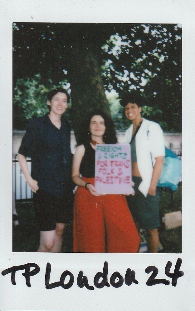 Three people are smiling in a park, holding a sign promoting rights for trans folk.