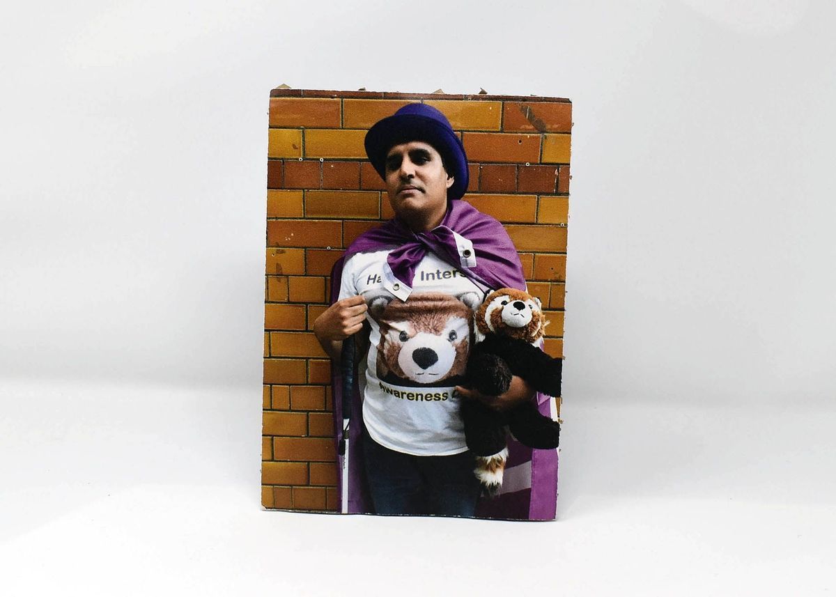 The person is wearing a purple flag and hat, holding a stuffed animal, standing against a tiled wall.