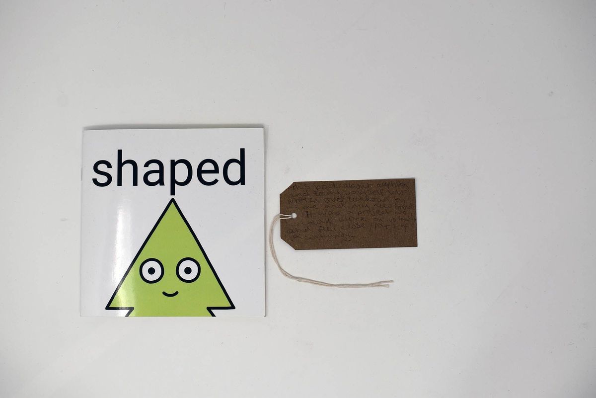 A small book entitled "shaped". The cover is white and shows a green triangular shaped cartoon character outlined in black. With a brown, handwritten luggage tag.