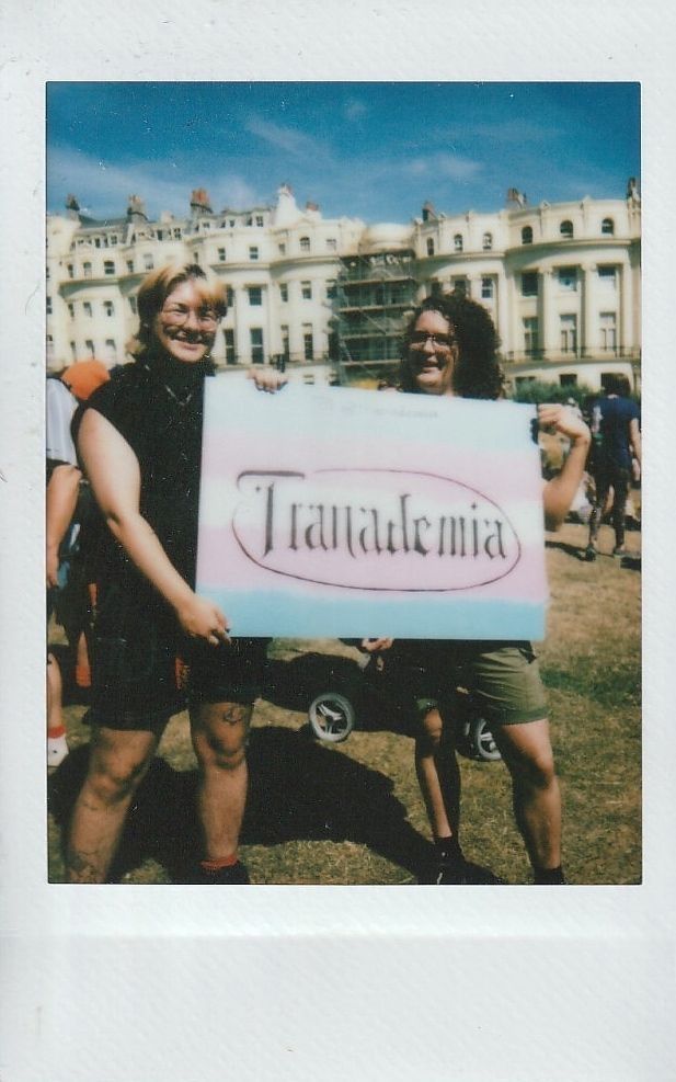 Two people standing outdoors hold a colorful sign labeled "Tranademia".