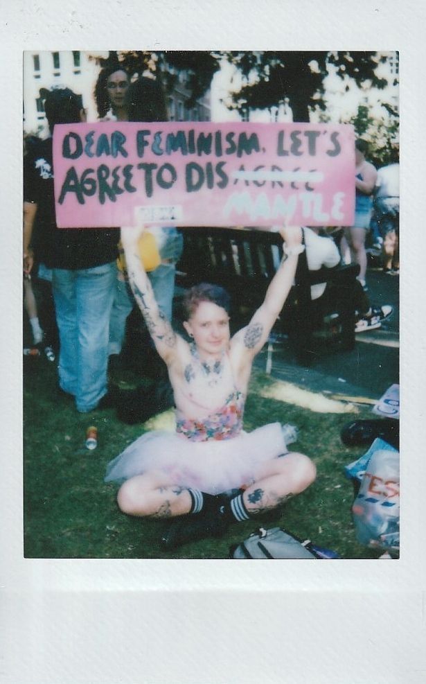 A person in a tutu holds a sign reading, "Dear feminism, let's agree to disMANTLE".