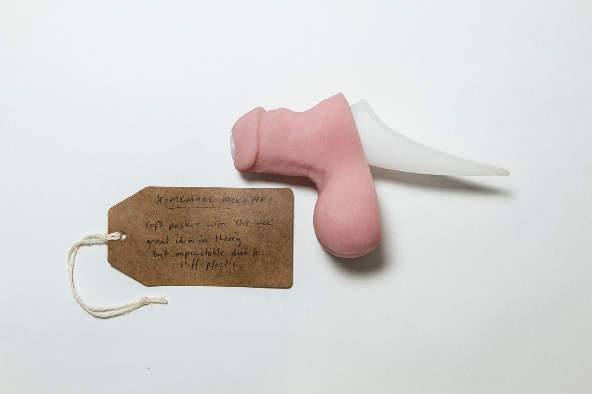 Prosthetic penis with "She Wee" attachment & tag; silicone, pink moulded penis & testicles with detailing of skin & hairs. Has been adapted with white plastic "Shee wee", trough and pipe have been inserted through penis base and head. Penis has flat base (back) across penis & testicles.