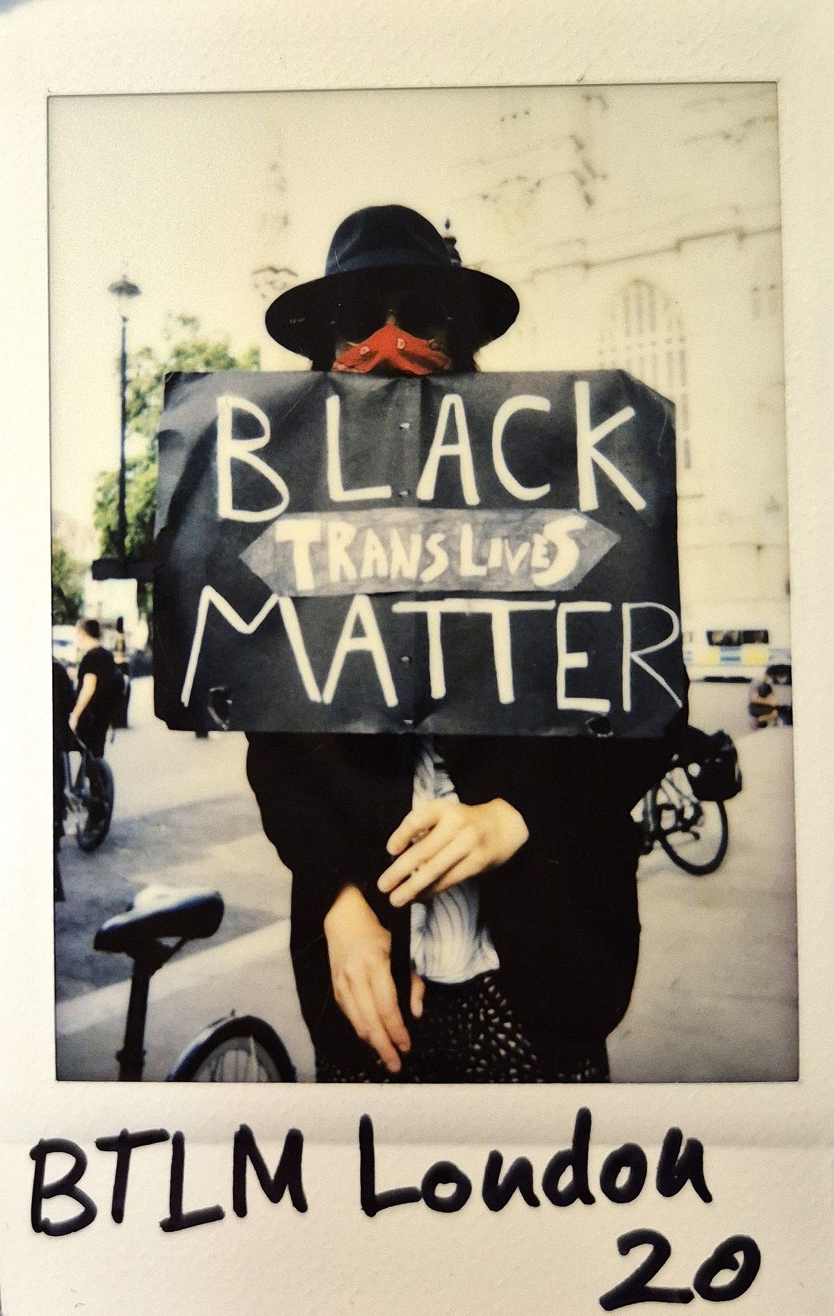 A person wearing a hat and mask holds a sign reading "Black Trans Lives Matter" at a protest.