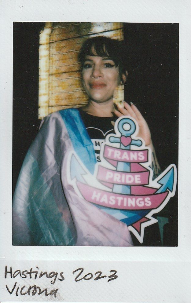 A person proudly holds a "Trans Pride Hastings" sign while wearing a flag at an event.