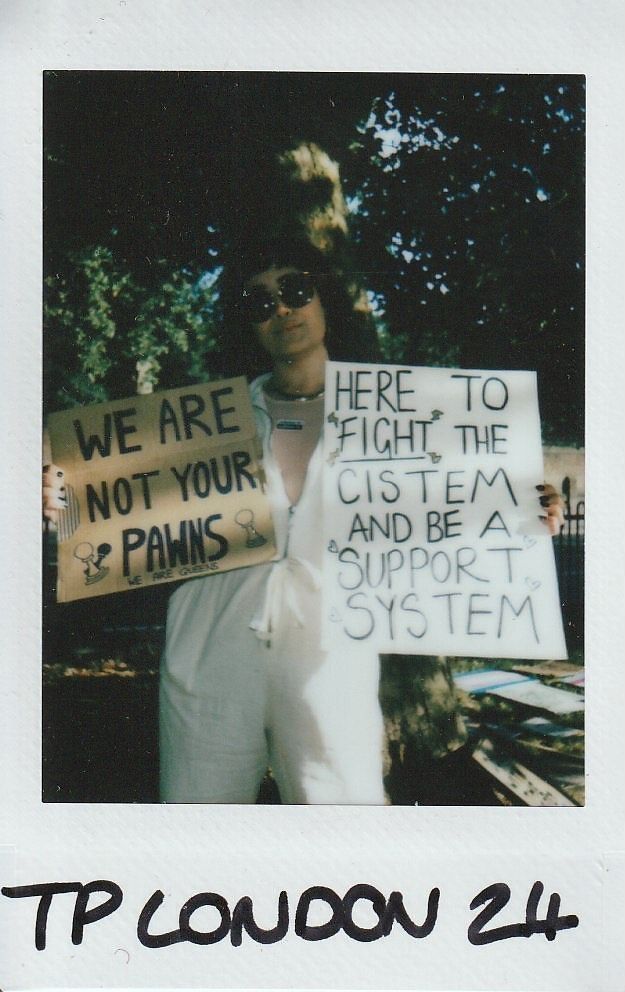 A person holds two protest signs while wearing sunglasses outdoors in a park setting with trees.