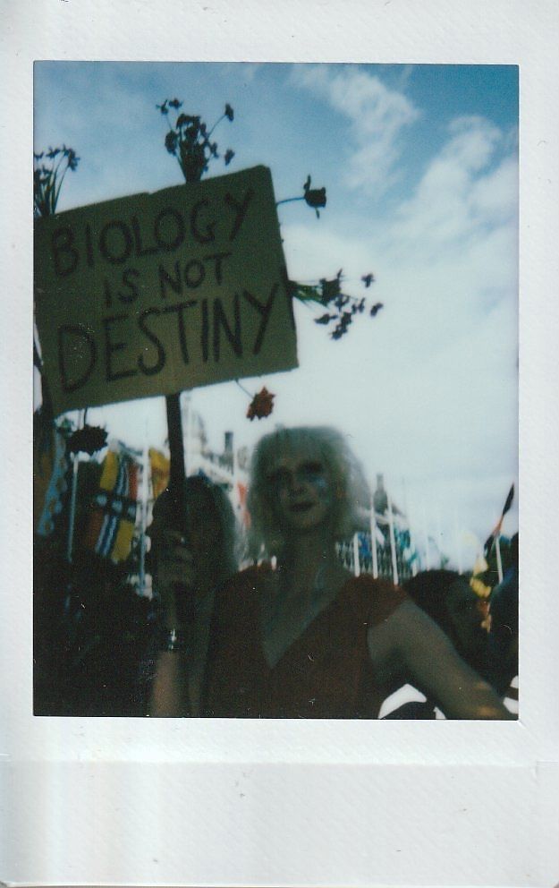 A person with blonde hair holds a protest sign that reads "Biology is not destiny".