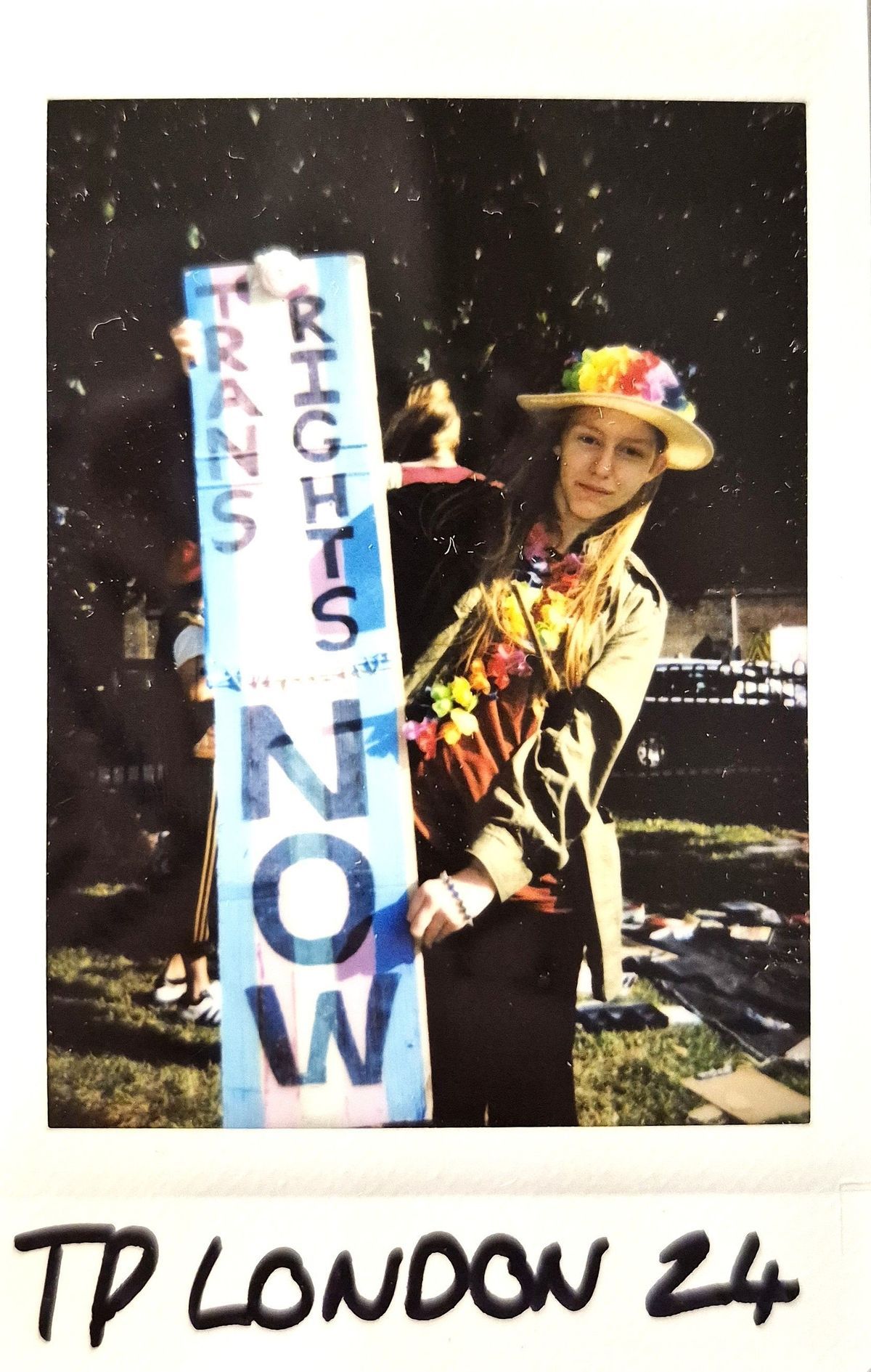 A person in a colorful hat holds a sign reading "Trans Rights Now" during an outdoor gathering.