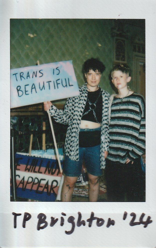 Two individuals stand together, one holding a sign that reads "Trans is Beautiful," expressing support and pride.