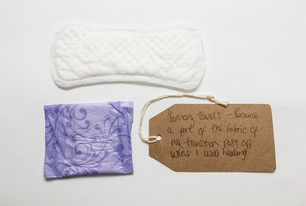 One white sanitary pad is laid lengthwise across the top of the photo. Directly underneath is a folded/ still packaged pad in light purple wrapping.