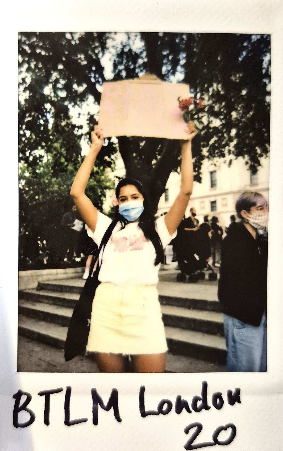 A person wearing a mask holds a sign overhead at a protest, standing on steps.