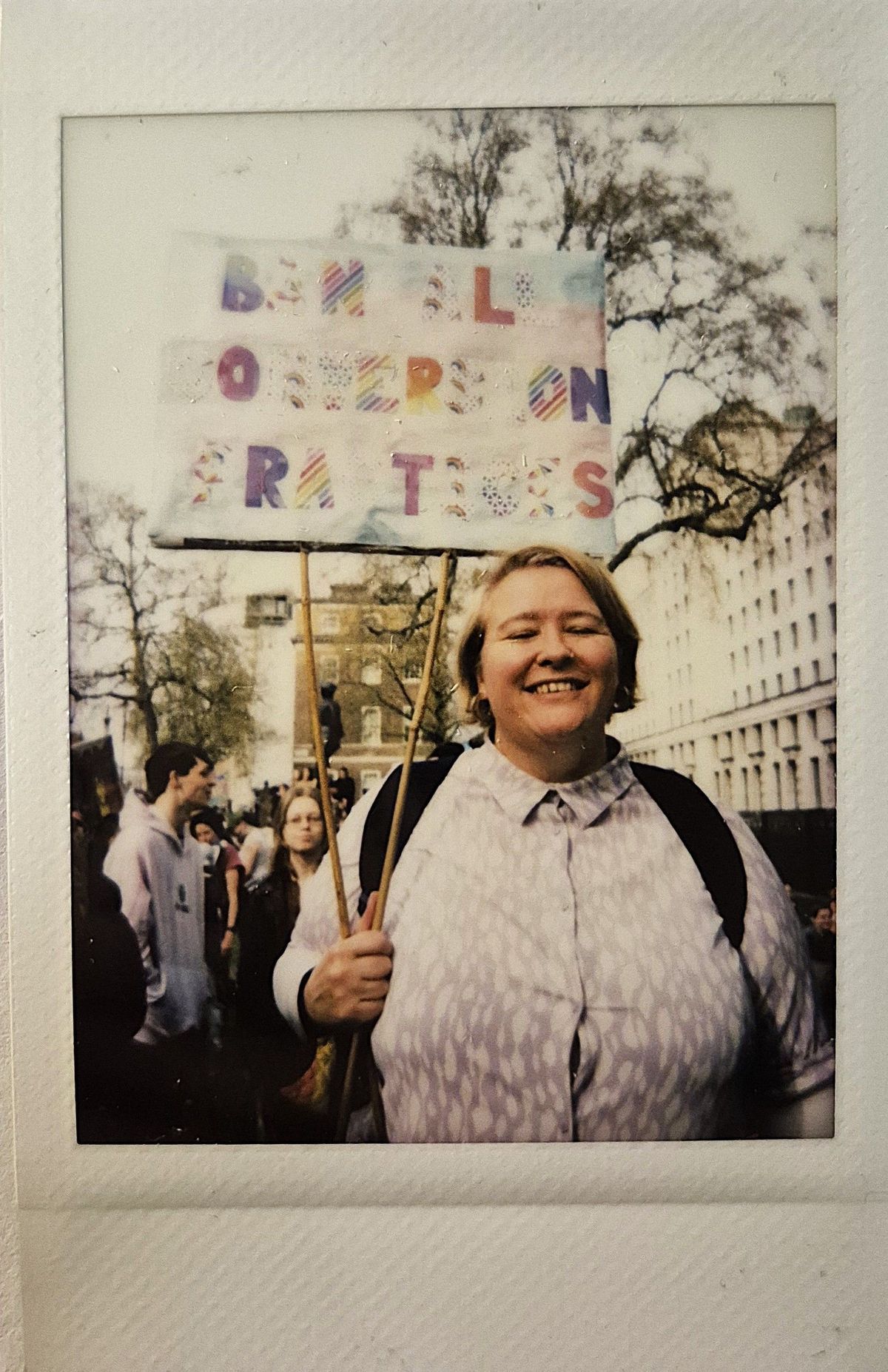 A smiling person holds a protest sign outdoors, surrounded by people and trees in the background.