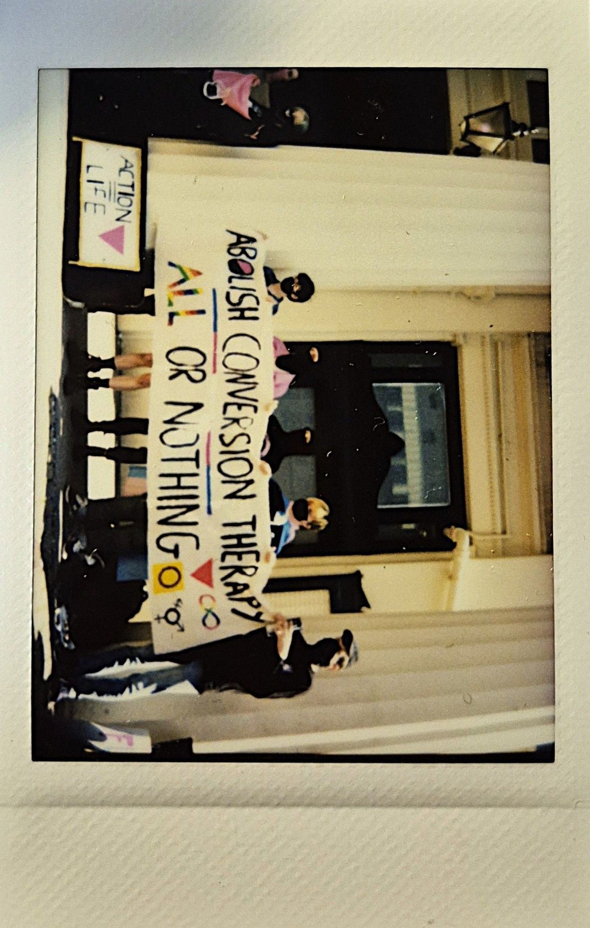 A group stands on steps holding a banner reading "Abolish conversion therapy, all or nothing."