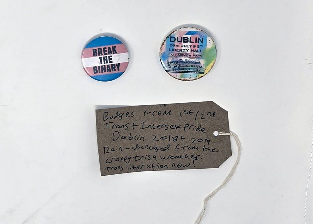 Tag reading "Badges from 1st/2nd Trans + Intersex Pride Dublin 2018 + 2019
Rain-damaged from the crappy Irish weather.
Trans liberation now!" Under the tag are two badges. Badge 1 reads "DUBLIN 28th July @ 2PM LIBERTY HALL TO FAIRVIEW PARK" and has a trans flag design background. Badge 2 reads "BREAK THE BINARY" and has a trans flag background.