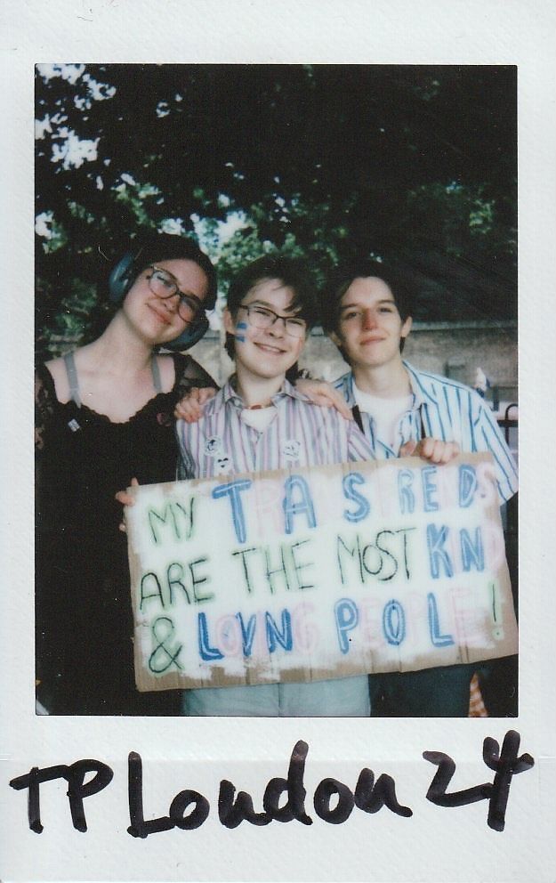 Three people stand together, smiling, holding a colorful sign with supportive text written on it.