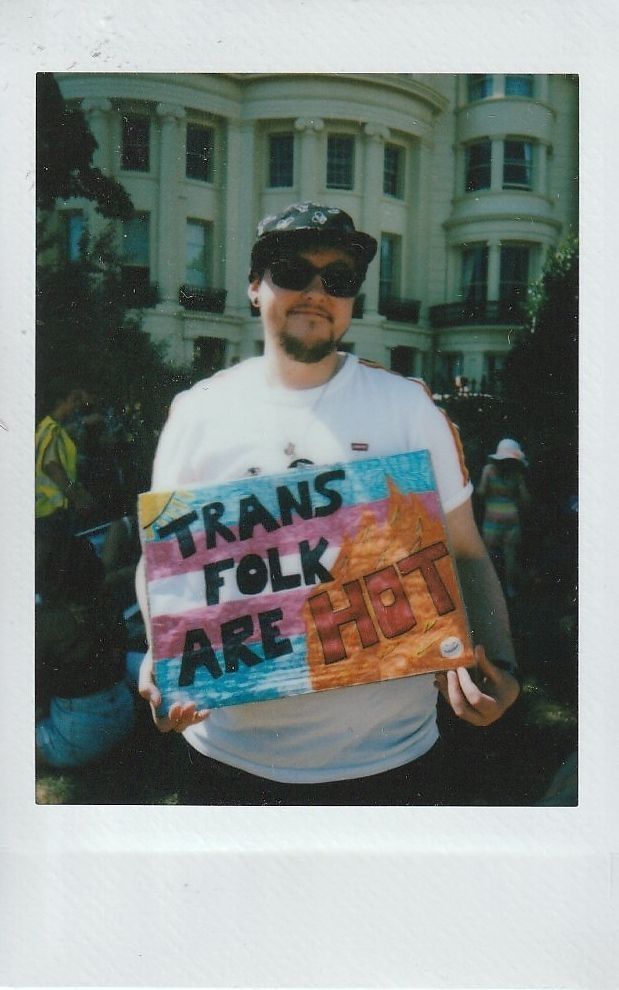 A person wearing sunglasses holds a colorful sign reading "Trans folk are hot" outside a large building.