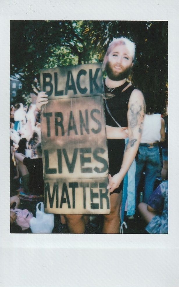 A person stands outside holding a large sign that reads "Black Trans Lives Matter" at a gathering.