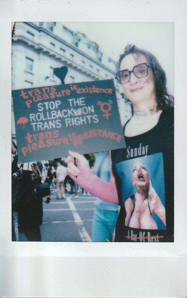 A person holds a sign which says "Trans Pleasure Is Existence Stop The Rollback On Trans Rights Trans Pleasure Is Resistence".