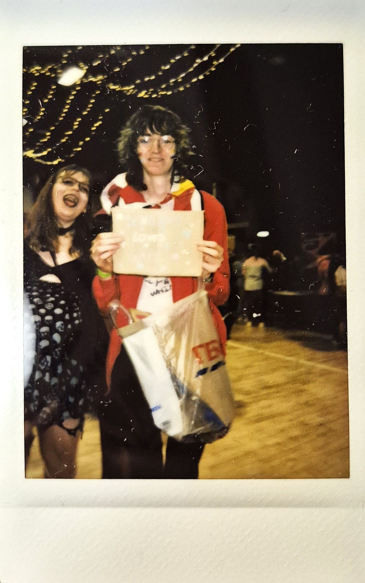 Two people smile cheerfully in a festive setting, with one holding a sign and bags.