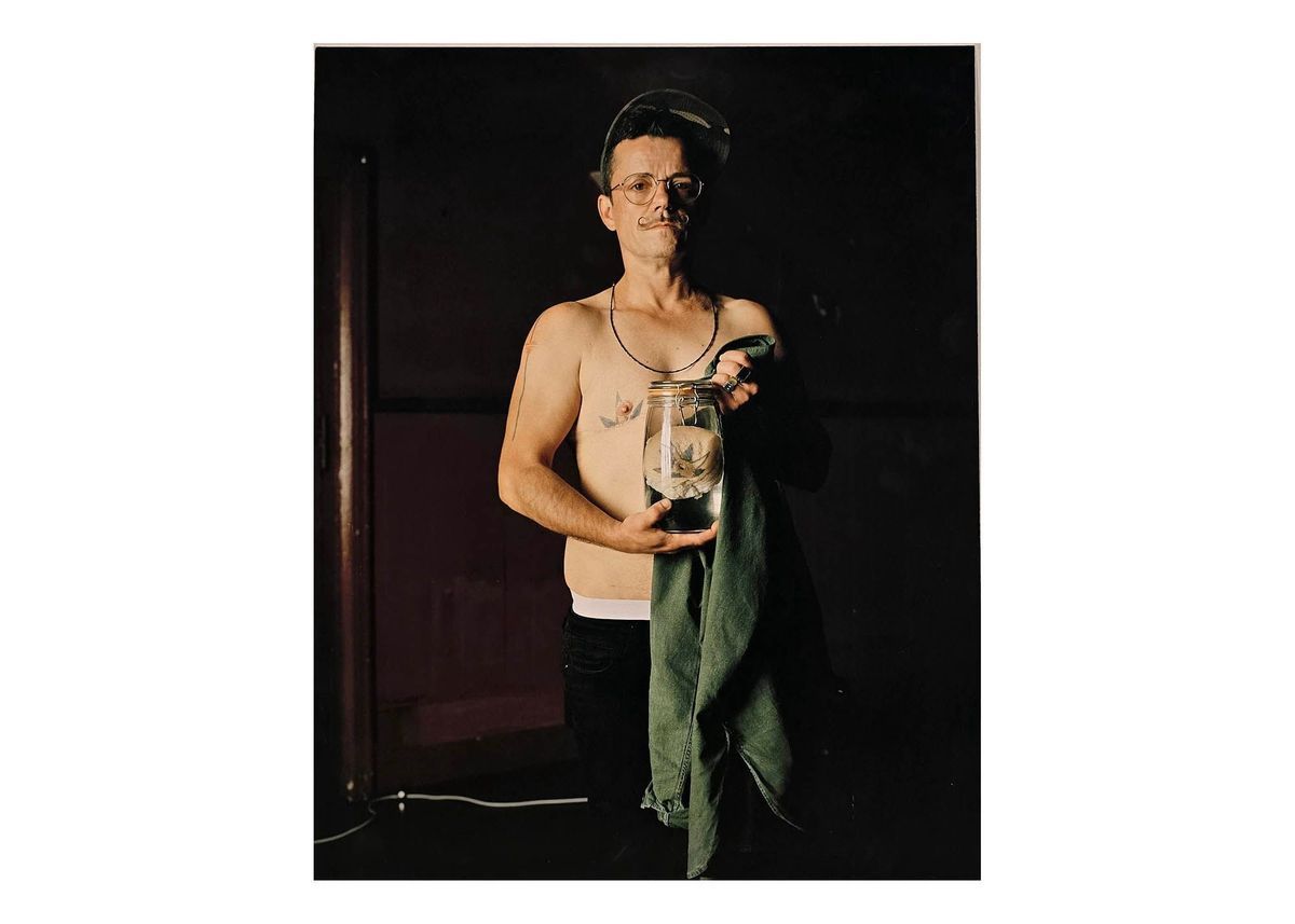 Colour photograph mounted onto metal back, unframed & unglazed; matt finish image of E-J Scott wearing green camouflage baseball cap, metal framed glasses, bare to waist, black trousers. Holding Kilner jar & green shirt. Jar contains human chest tissue. Image taken against dark background.