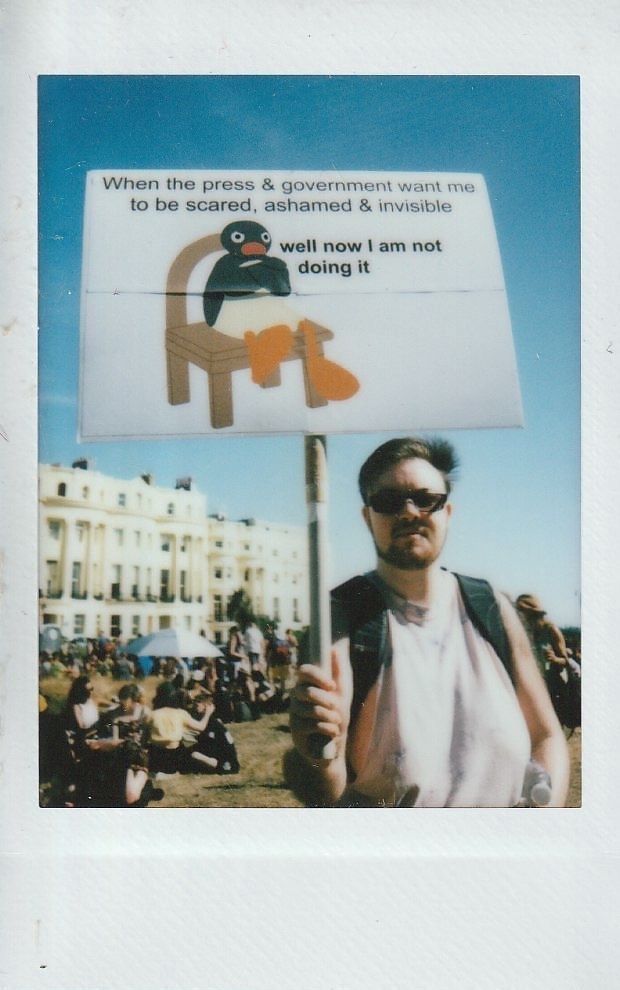 A person stands holding a protest sign which says "When the press & government want me to be scared, ashamed & visible" with an image of Pingu the penguin sat on a chair which then reads "well now I am not doing it".