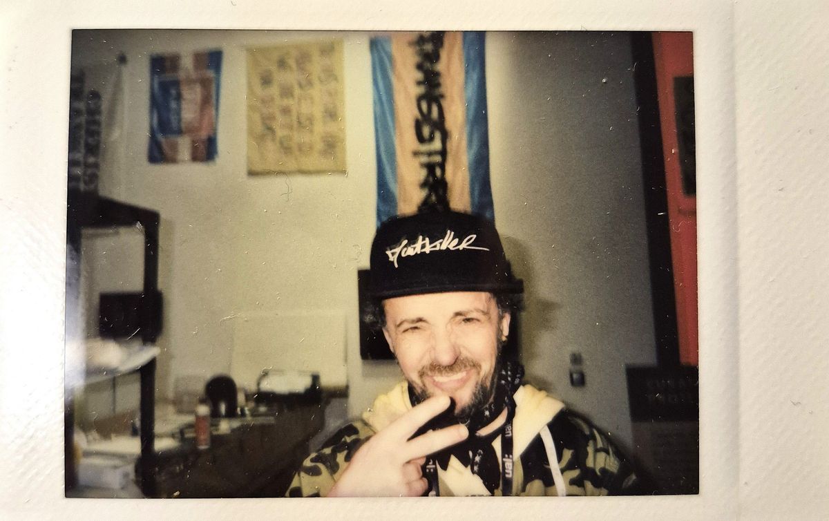 A smiling person wearing a cap flashes a peace sign while posing indoors with flags behind them.