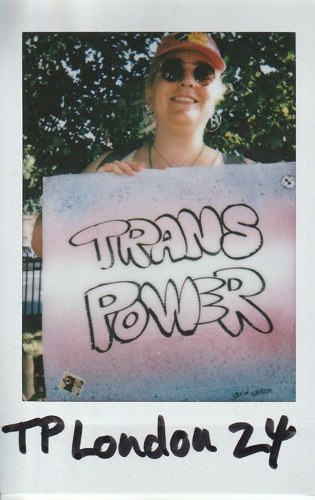 A person wearing sunglasses and a hat holds a sign reading "Trans Power" with trees in the background