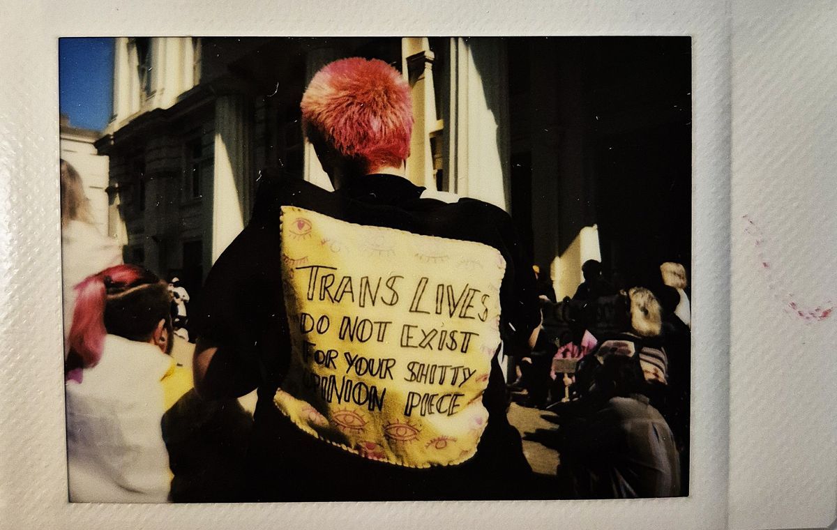A person with pink hair wears a jacket displaying a message on the back which says "TRANS LIVES DO NOT EXIST FOR YOUR SHITTY OPINION PIECE".