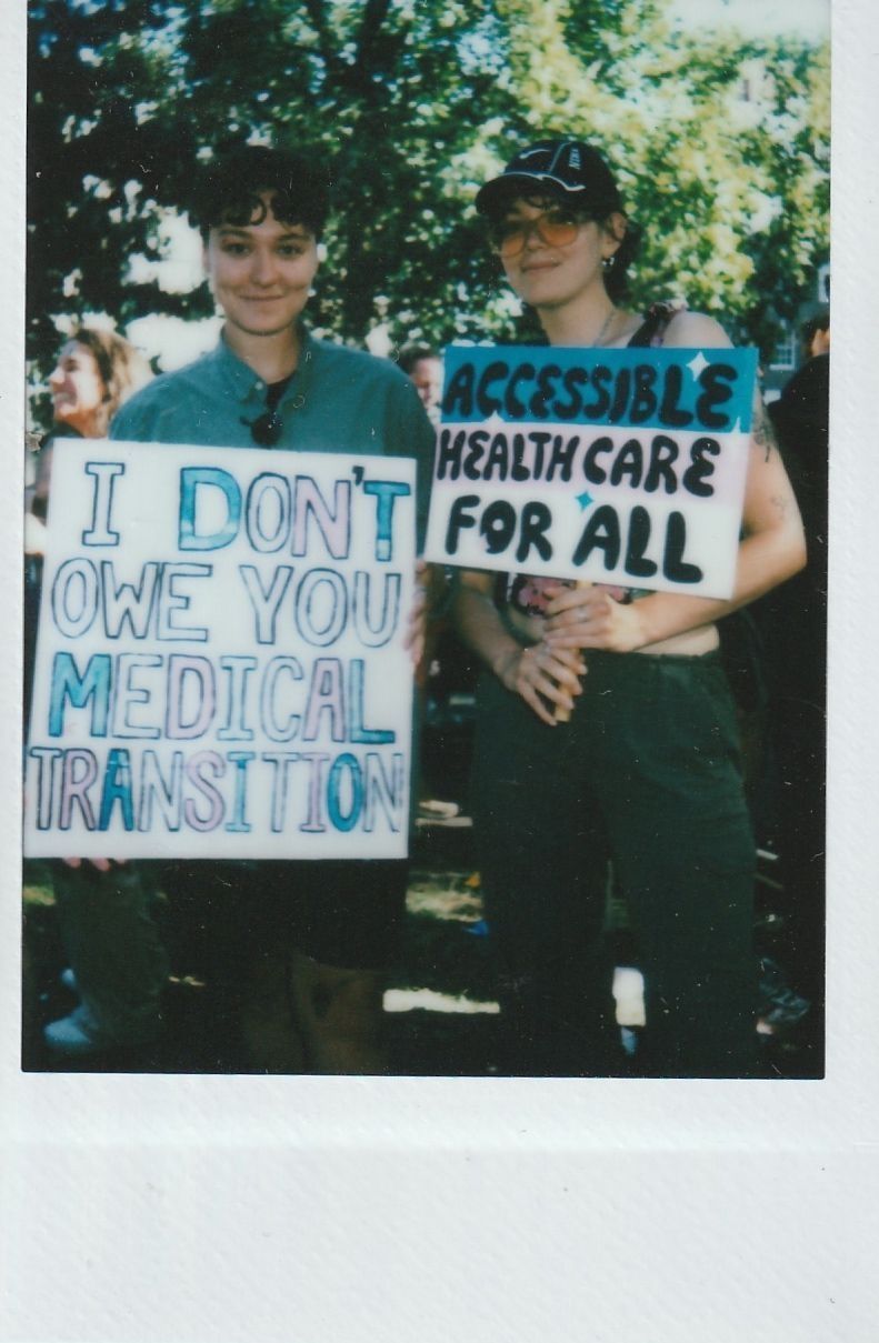 Two people are holding signs, one which says "I DON'T OWE YOU A MEDICAL TRANSITION" and the other "ACCESSIBLE HEALTHCARE FOR ALL".