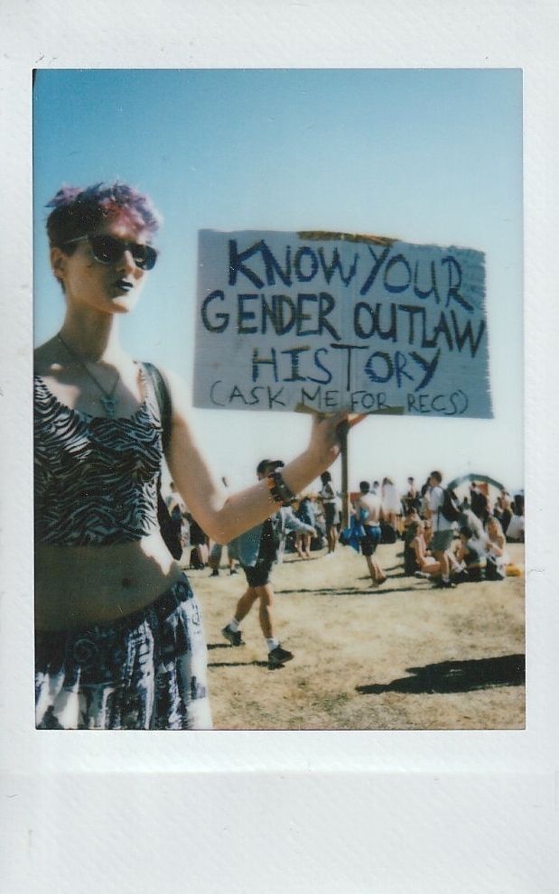 A person holds a sign reading "Know Your Gender Outlaw History" at an outdoor event.