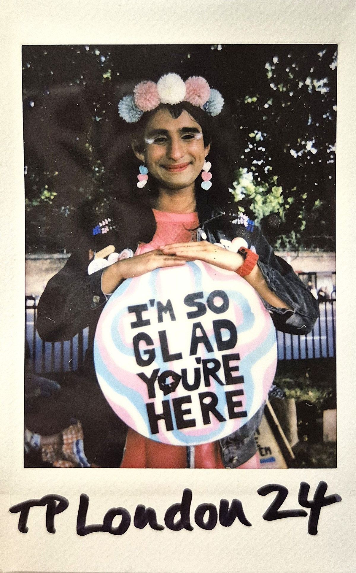 A person smiles warmly, wearing colorful pompom accessories and holding a sign that says, "I'm so glad you are here"