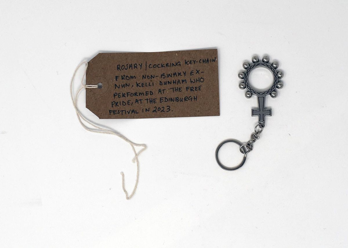 A silver rosary cock ring pendant. Next to it on the left is a brown tag with writing on it.