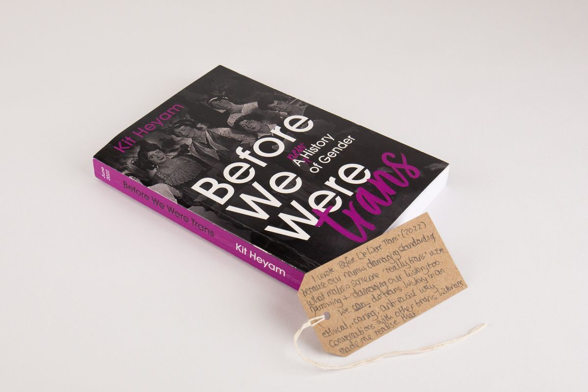 Book entitled "Before We Were trans: A NEW History of Gender" by Kit Heyam. The books spine is magenta and the cover features an old black and white photo of some formally dressed people. With a brown, handwritten luggage tag.