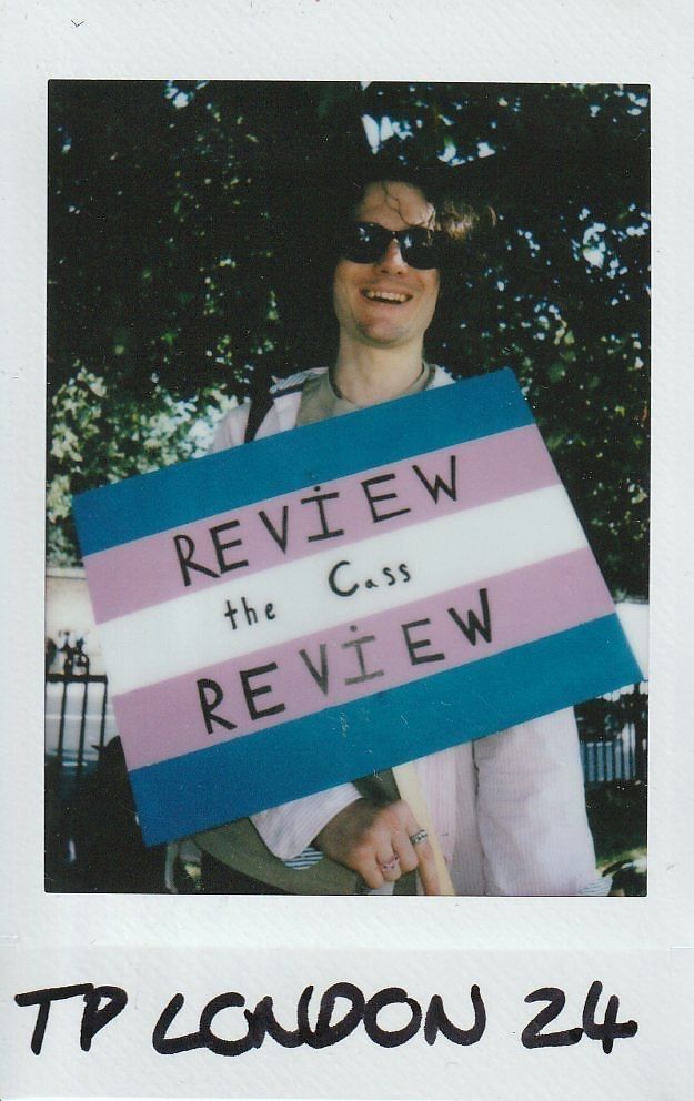 A person holding a colorful protest sign stands outside, wearing sunglasses and smiling near the trees.