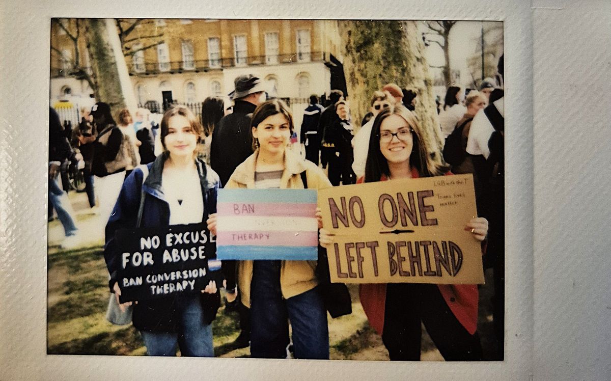 Three individuals stand holding signs advocating for banning conversion therapy and inclusivity during a protest. One sign says "NO EXCUSE FOR ABUSE BAN CONVERSION THERAPY", one says "BAN CONVERSION THERAPY" and the third says "NO ONE LEFT BEHIND".