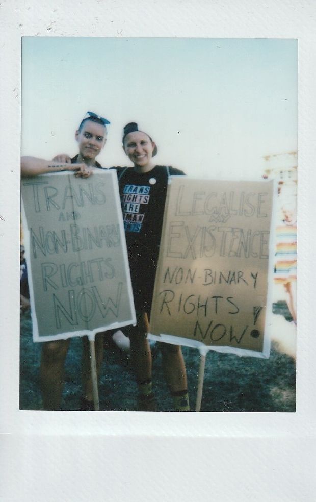 Two people hold signs which say "Trans and Non-binary Rights Now" and "Legalise My Existence Non-binary Rights Now".