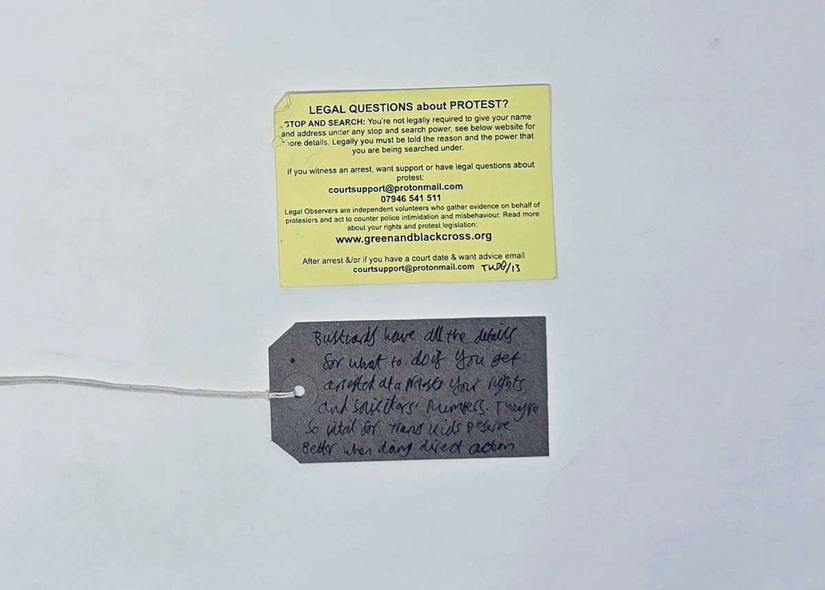 A yellow "Bust Card" from Green and Black Cross detailing a persons rights around Stop and Search and contact details for further advice. With a brown, handwritten luggage tag.