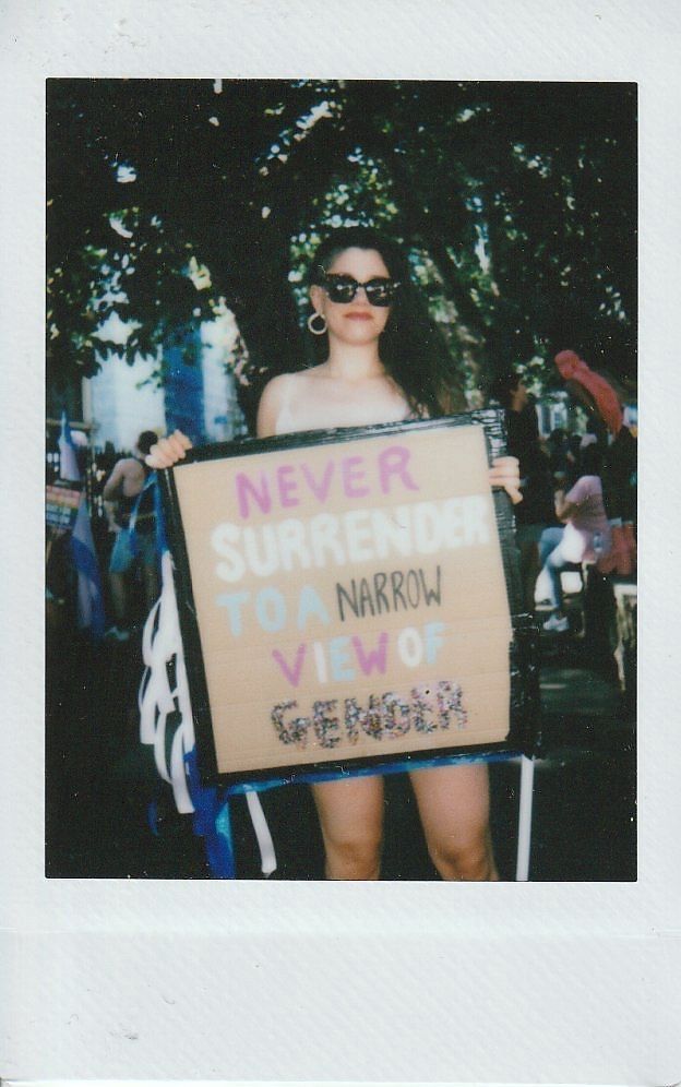 A person is holding a sign that reads "Never Surrender to a Narrow View of Gender".
