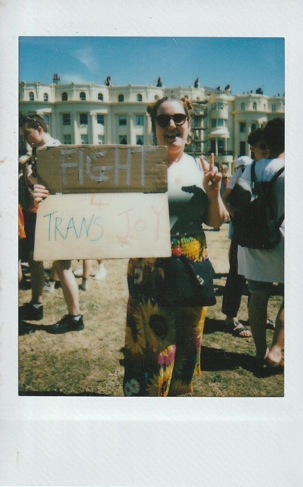 A person wearing sunglasses holds a sign saying "Fight for Trans Joy" in a sunny outdoor gathering.