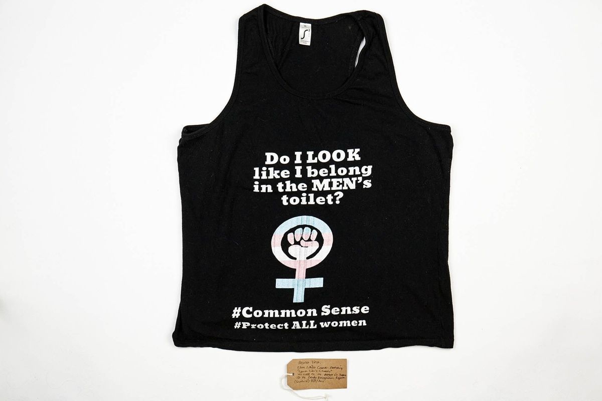 A black vest top depicting the feminist resistance symbol in the trans colours, stylised with a raised fist and the text "Do I LOOK like I belong in the MEN's toilet" "#CommonSense""#ProtectALLwomen". Also, a brown, handwritten luggage tag.