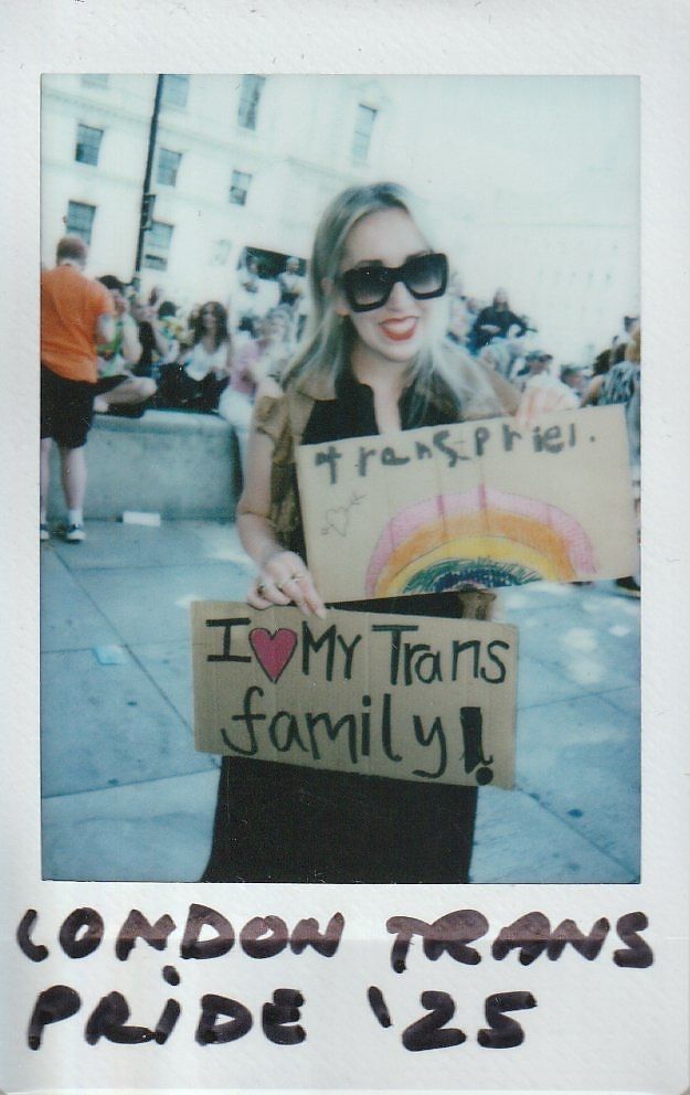 A person smiles, holding signs supporting the trans community, one says "Trans Pride" with a rainbow drawn on and the other says "I <3 My Trans Family".