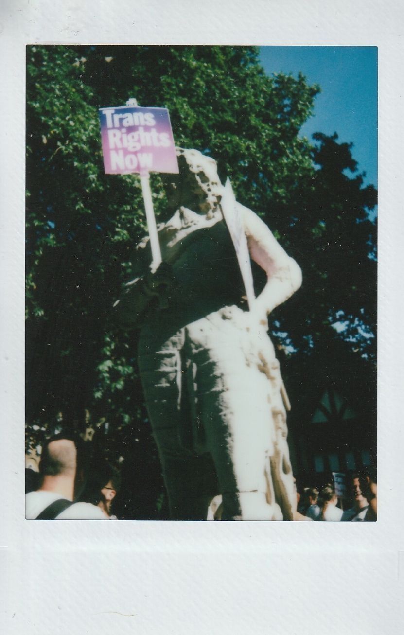 A statue holds a sign reading "Trans Rights Now" at a gathering outdoors under a bright blue sky