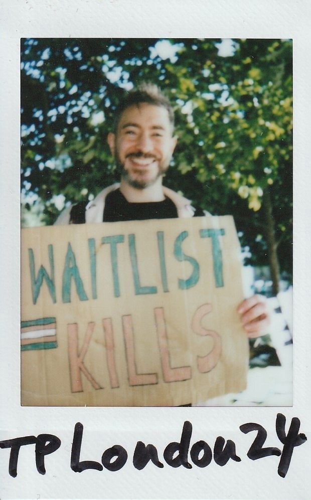 A smiling person holds a sign reading "WAITLIST KILLS" in a sunny park setting.