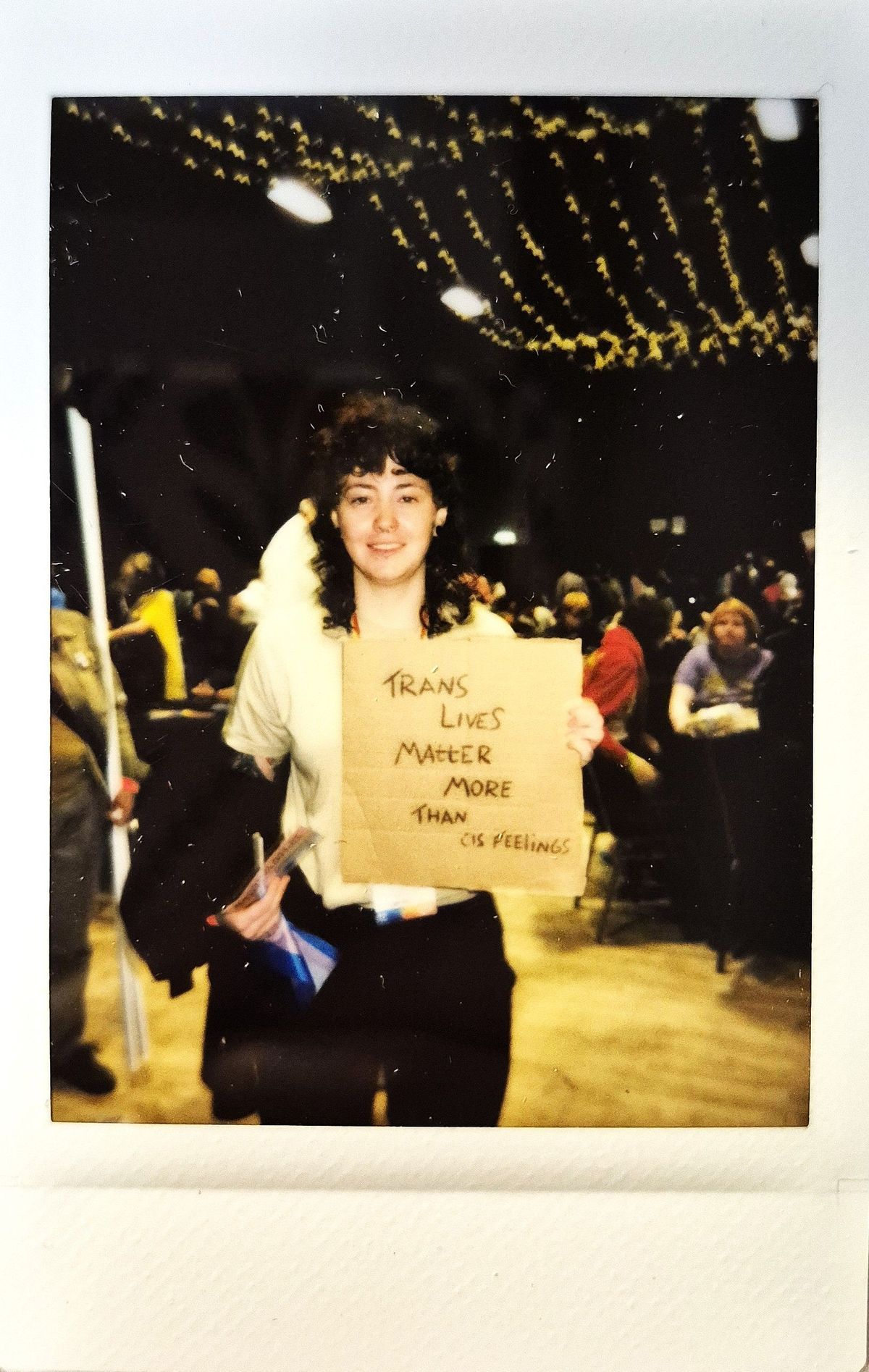 A person holds a sign reading "Trans lives matter more than cis feelings" at a crowded event.