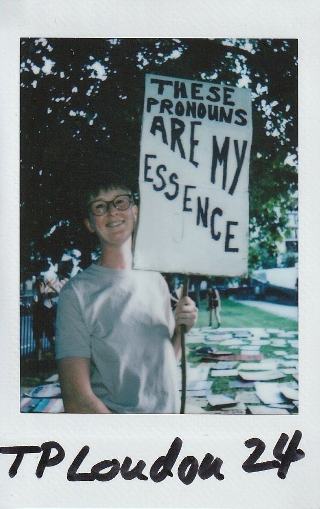 A person holds a sign saying, "These pronouns are my essence," while standing outdoors under trees