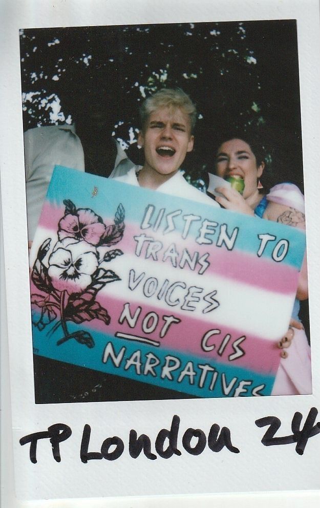 Two individuals hold a colorful sign advocating for listening to trans voices over cis narratives.
