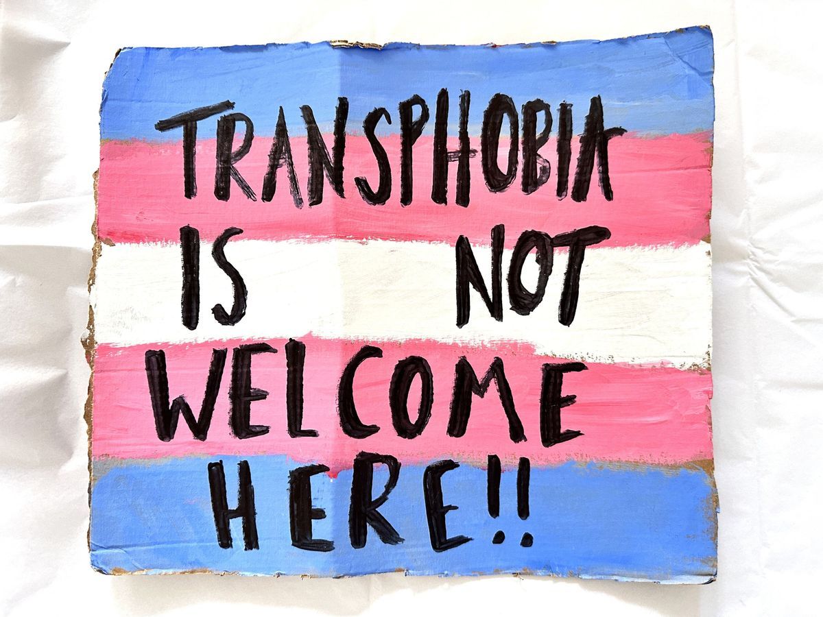 The sign displays "Transphobia is not welcome here" on a flag background.