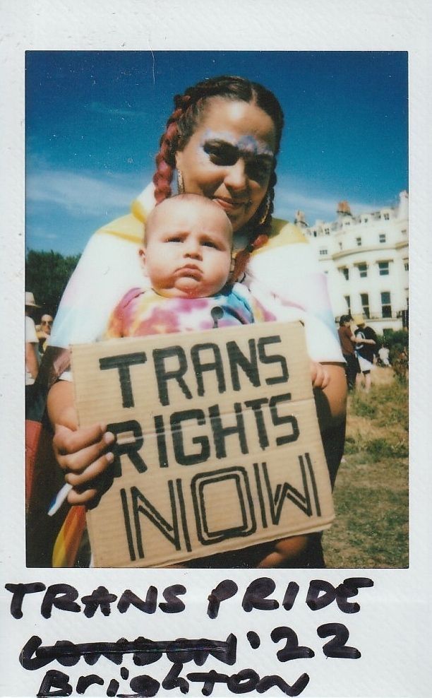 A person with colorful makeup holds a baby and a "Trans Rights Now".