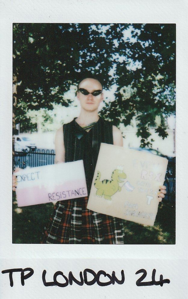 A person in sunglasses holds two signs, "Expect Resistance" and a dinosaur drawing, outdoors.
